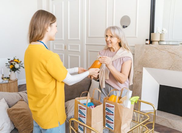 A senior woman receiving groceries from a young caregiver in a cozy living room setting.